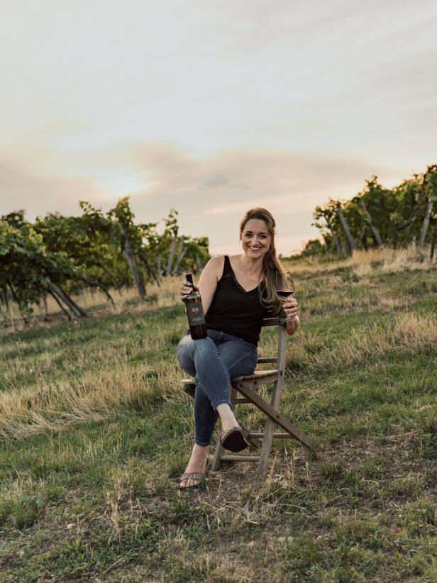 Christin Netzl sitzt mit einer Flasche Wein und einem Glas in einem Weinberg.