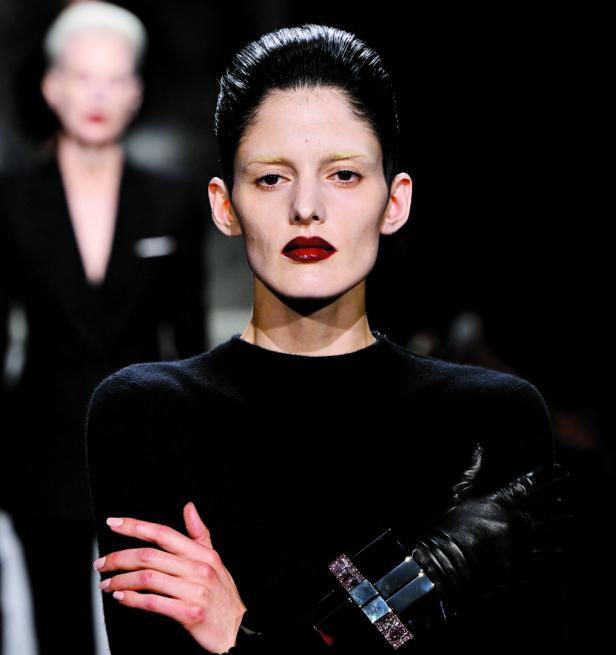 Eine Frau mit dunklem Lippenstift und einem schwarzen Pullover posiert mit verschränkten Armen.