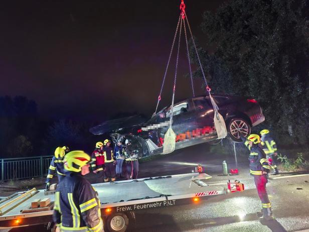 Mittels Krank wird ein Pkw nach dem frontalen Zusammenstoß auf eine Ladevorrichtung zum Abtransport gehieft. Mehrere Feuerwehrmänner stehen im Dunklen um die Fahrzeuge herum.