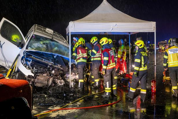 Mehrere Menschen in Feuerwehrkleidung stehen Nachts und einem weißen Zelt neben einem demolierten Auto.