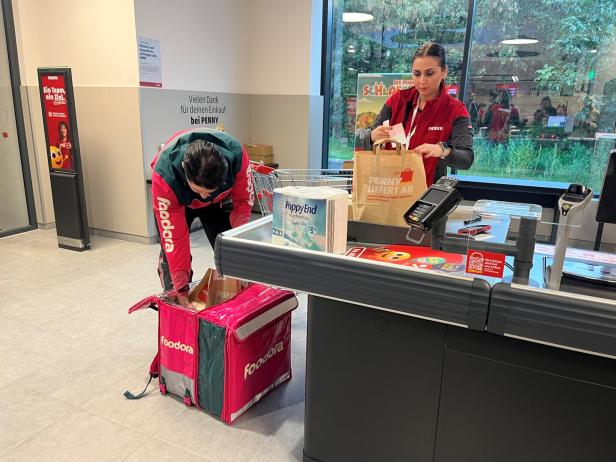 Eine Penny-Mitarbeiterin packt Einkäufe ein, während ein Foodora-Kurier daneben seinen Rucksack befüllt.
