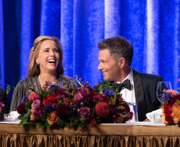 Téa Leoni und Tim Daly sitzen lachend hinter einem Tisch mit bunten Blumenarrangements.