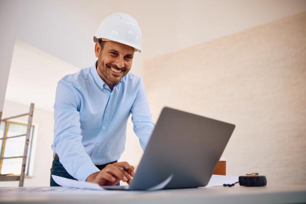 Ein Mann mit einem Helm arbeitet an einem Laptop auf einer Baustelle..