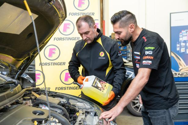 Zwei Männer prüfen in einer Autowerkstatt den Motorraum eines Fahrzeugs und füllen gelbe Flüssigkeit ein.