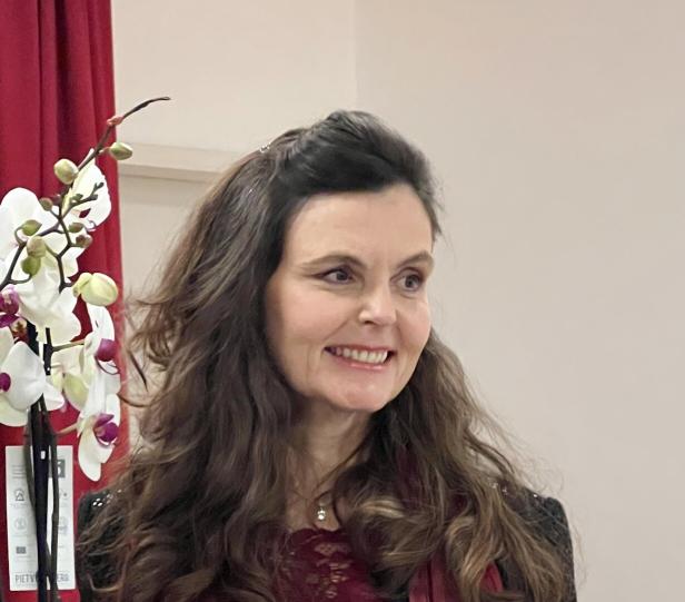 Eine lächelnde Frau mit langen, braunen Haaren steht neben einer weißen Orchidee.