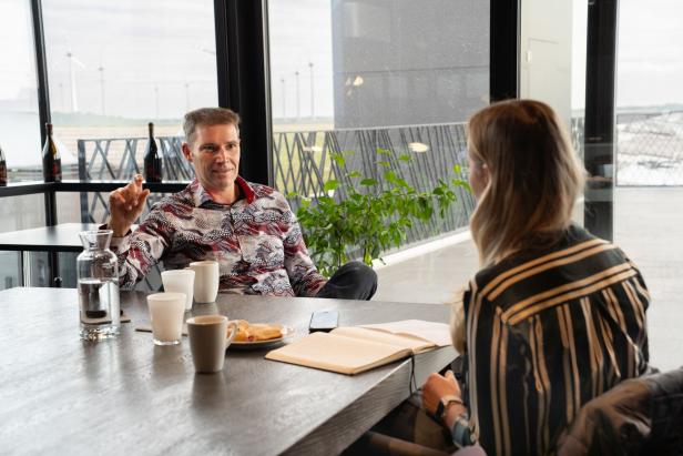 Zwei Personen unterhalten sich an einem Tisch mit Getränken, Gebäck und einem offenen Notizbuch in einem modernen Café.
