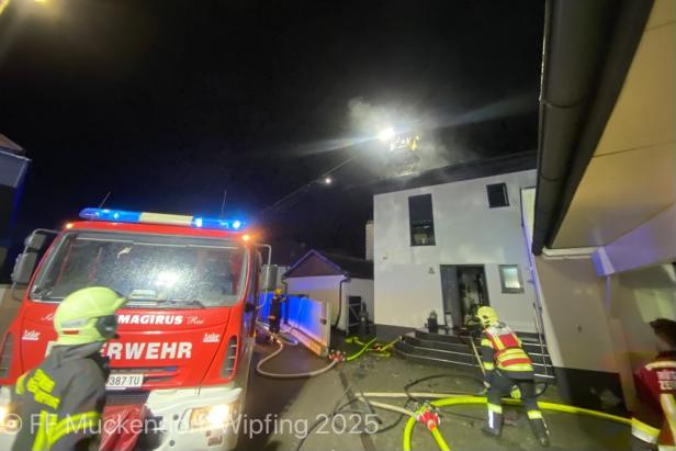 Ein Feuerwehrauto steht in der Nacht vor einem brennenden Haus in Tulln, zwei Feuerwehrmänner stehen mit dem Rücken zur Kamera.