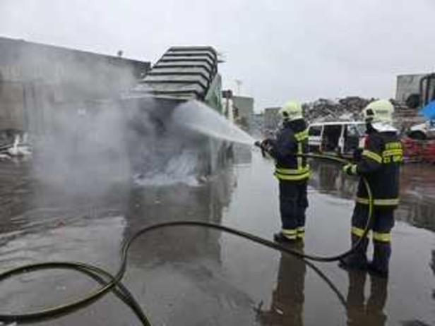Containerbrand Zwei Feuerwehrleute löschen mit einem Wasserschlauch ein qualmendes Feuer auf einem Industriegelände.