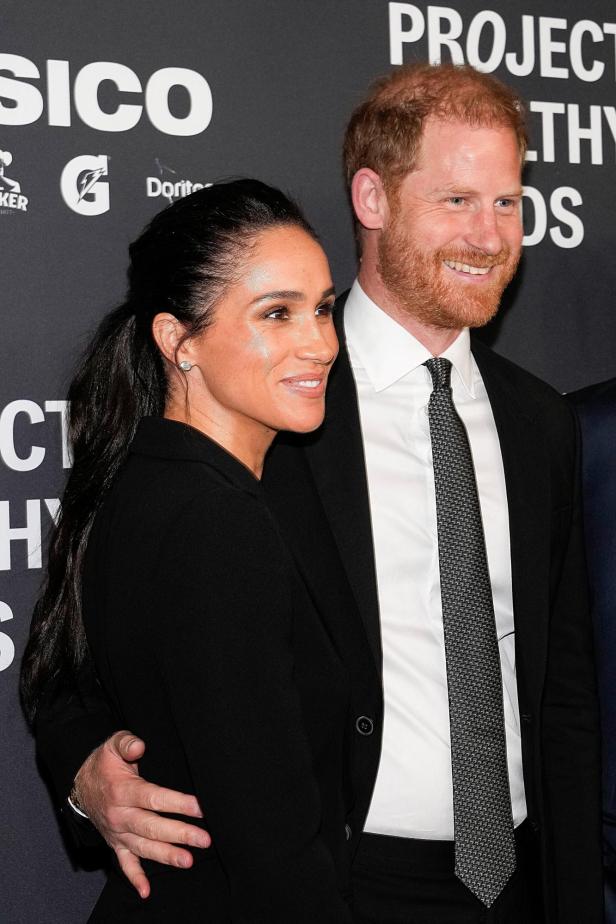 Meghan und Prinz Harry lächeln auf einer Veranstaltung vor einem dunklen Hintergrund.