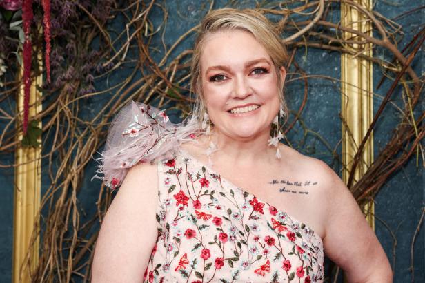 Eine lächelnde Frau mit blondem Haar, Colleen Hoover, trägt ein geblümtes, schulterfreies Kleid und auffällige Ohrringe vor einem Hintergrund mit Ästen.