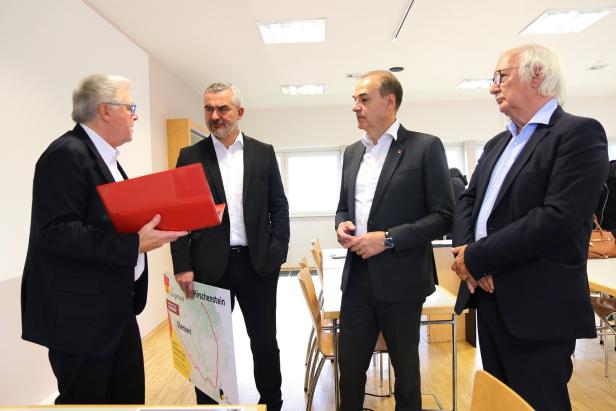 Vier Männer in Anzügen besprechen etwas, einer hält eine rote Mappe und eine Karte von Oberwart und Hirschenstein.