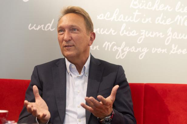 Ein Mann im Anzug gestikuliert vor einer Wand mit Schriftzügen.