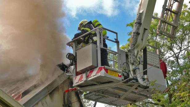 Ein Feuerwehrmann steht im Korb einer Drehleiter, vor ihm eine dichte Rauchschwade, die aus dem Dach aufsteigt.
