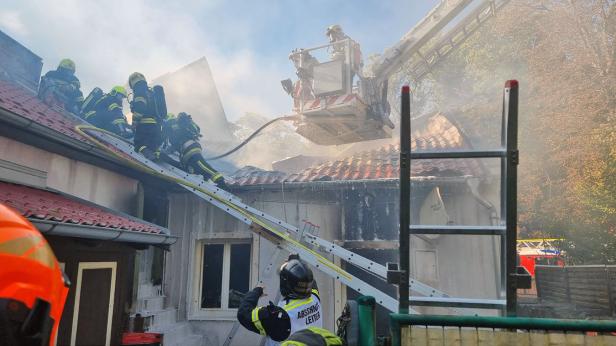 Eine Aufnahme vom Brandort, mehrere Feuerwehrleute sind mit den Löscharbeiten beschäftigt, Rauch steigt auf.