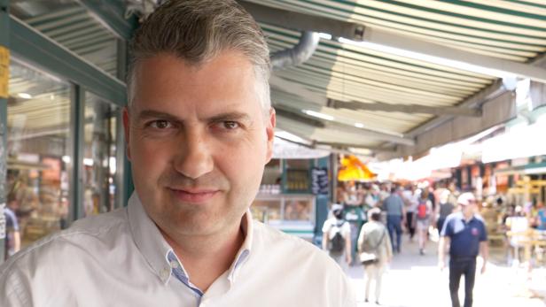 Ein Mann mit grauen Haaren und weißem Hemd steht auf einem belebten Markt mit vielen Menschen im Hintergrund.