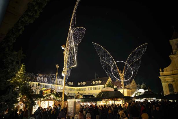 Zwei große Engel aus Lichterketten über einem belebten Weihnachtsmarkt bei Nacht, umgeben von historischen Gebäuden.