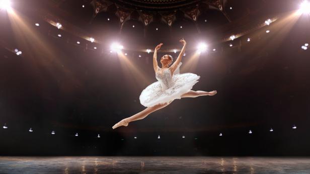 Ballerina in weißem Tutu springt mit ausgestreckten Armen und Beinen auf einer Bühne mit Scheinwerfern im Hintergrund.