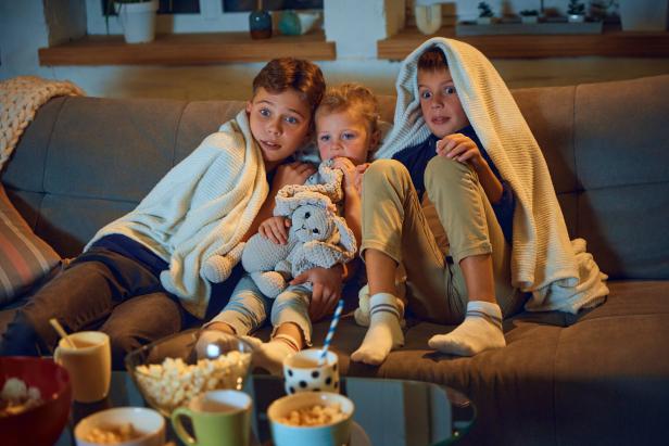 Emotional children, boys and little girl sitting on couch at home and watching tv, horror movie. Hiding under blanket Kinder schauen gemeinsam einen Film und fürchten sich.