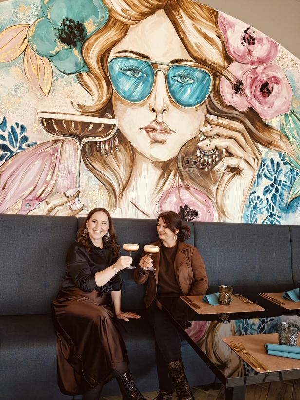 Zwei Frauen stoßen mit Cocktails an, im Hintergrund eine bunte Wandmalerei einer Frau mit blauer Sonnenbrille.