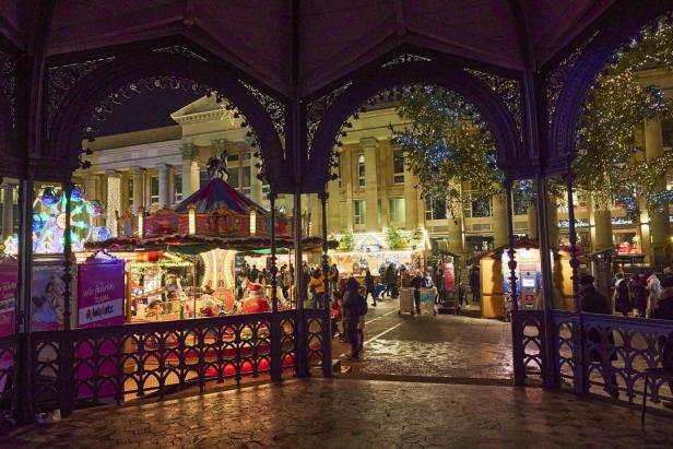 Blick durch ein verziertes Holzpavillon auf einen beleuchteten Weihnachtsmarkt mit Karussell und Menschen vor einem historischen Gebäude bei Nacht.
