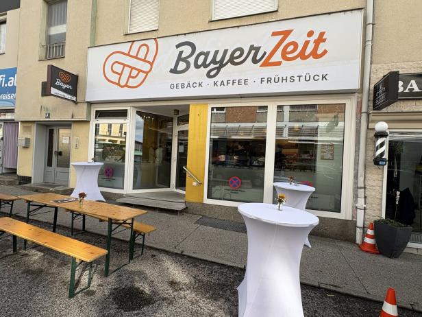 Vor einer Bäckerei mit dem Namen „BayerZeit“ stehen Holzbänke und mit weißen Tüchern bedeckte Stehtische mit kleinen Blumenvasen.