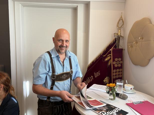 Ein Mann in Tracht steht lächelnd an einem Tisch mit Unterlagen und einer Fahne.
