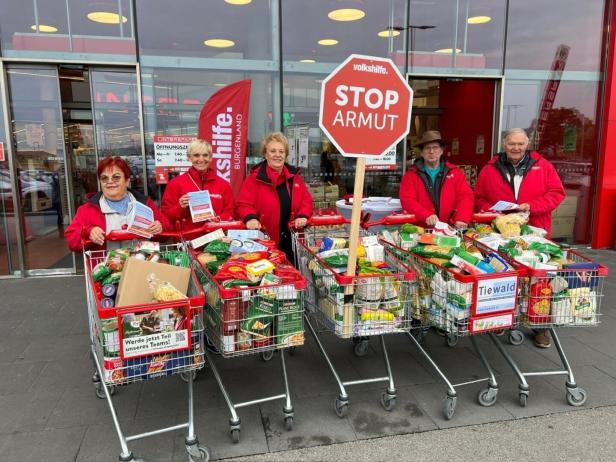 Vor einem Supermarkt stehen Helfer der Volkshilfe mit Einkaufswagen voller Lebensmittel und einem "STOP Armut"-Schild.