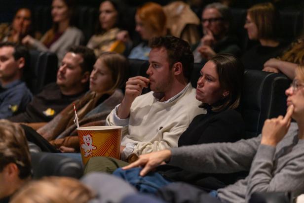 Mehrere Menschen sitzen nebeneinander in einem Kinosaal und schauen konzentriert nach vorne, während einer Popcorn isst.