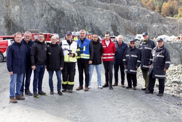 Eine Gruppe von Männern in Einsatzkleidung und Jacken steht vor Felswänden und Einsatzfahrzeugen im Freien zusammen.