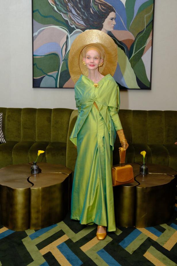 Eine elegant gekleidete Frau in grünem Kleid und großem goldenen Hut steht vor einem Gemälde auf einem grünen Sofa.