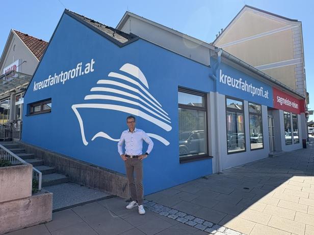 Ein Mann steht vor einem Gebäude mit der Aufschrift "kreuzfahrtprofi.at" und einem Schiffslogo.