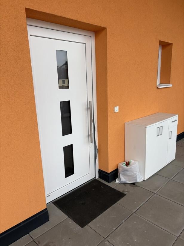 Weiße Haustür mit drei schmalen Fenstern, daneben ein weißer Schrank und ein Müllsack auf grauen Fliesen vor orangefarbener Wand.