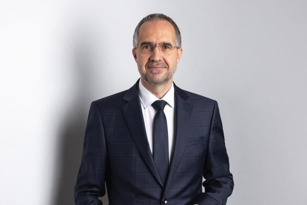 Vivatis Chef Gerald Hackl Ein Mann mit Brille und Anzug steht vor einer hellen Wand.