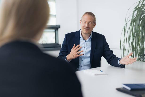 Mann im Anzug sitzt an einem Tisch, gestikuliert mit beiden Händen, gegenüber eine Frau mit blondem Haar.