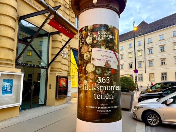 Werbesäule mit Plakat für Kunsthistorisches Museum, zeigt Blumenmuster und Text 'Gutschein Jahreskarte', '365 Glücksmomente teilen', 'Mit Ihrer Jahreskarte selbst', 'www.khm.at' vor Gebäude und parkenden Autos.