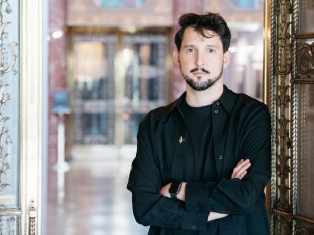 Ein Mann mit Bart in einem schwarzen Anzug steht mit verschränkten Händen in einem Türrahmen und schaut ernst in die Kamera.