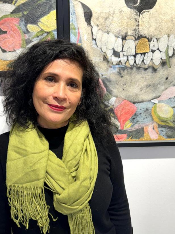 Eine Frau mit dunklen Haaren und grünem Schal steht vor einem Kunstwerk, das einen Totenschädel mit bunten Elementen zeigt.