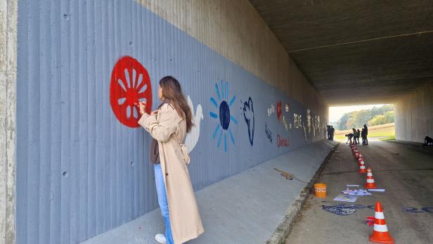 Eine Frau bemalt die Wand einer Unterführung mit bunten Symbolen, während weitere Personen im Hintergrund arbeiten.
