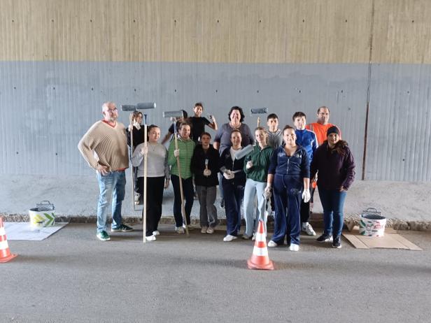 Eine Gruppe von Menschen steht mit Farbrollen und Pinseln vor einer bemalten Betonwand unter einer Brücke.