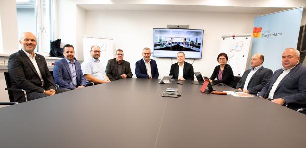 Neun Personen sitzen an einem Konferenztisch, im Hintergrund sind ein Bildschirm und Präsentationsmaterial zu sehen.