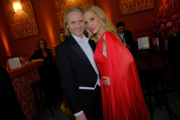 Ein elegant gekleidetes Paar posiert eng umschlungen in einem festlich dekorierten Raum mit roten Wänden.