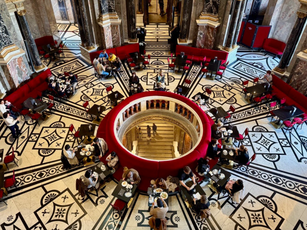 Blick von oben auf eine Cafeteria mit rundem Treppenaufgang in der Mitte und schwarz-weiß gemustertem Boden, umgeben von roten Sitzbänken und Tischen mit Gästen.