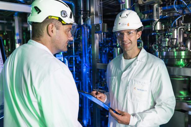 Zwei Personen in weißen Schutzanzügen und Helmen stehen in einer Industrieanlage, eine hält ein Klemmbrett und einen Stift.