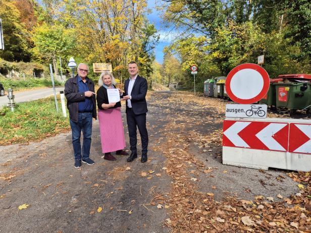 Drei Personen stehen auf einer herbstlichen Straße, halten gemeinsam ein Dokument und lächeln in die Kamera.