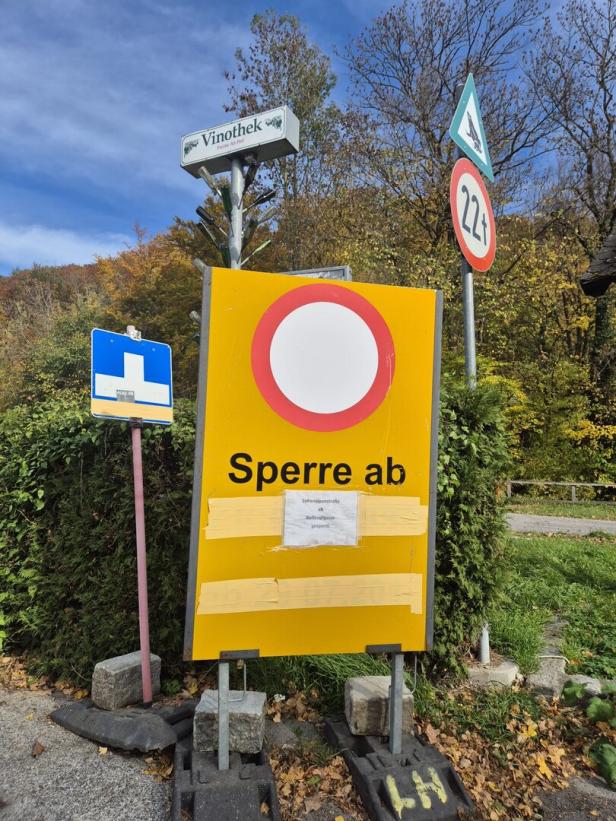 Straßensperre mit Hinweisschild „Sperre ab“, umgeben von weiteren Verkehrszeichen und herbstlicher Vegetation.