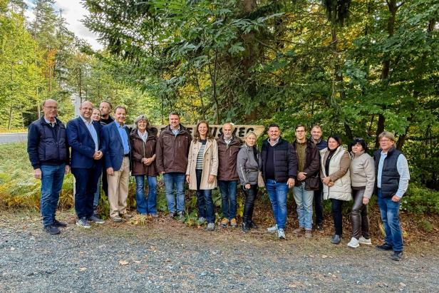 Eine Gruppe von Menschen steht nebeneinander im Freien vor einem Wald und einem Holzschild.