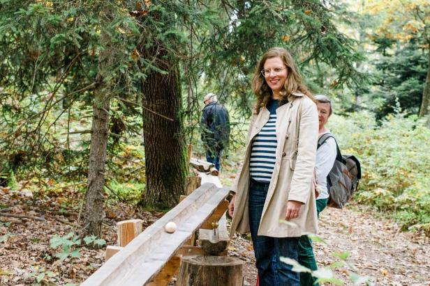 Eine Frau mit Brille steht lächelnd an einer Holzkugelbahn im Wald, umgeben von Bäumen und Laub.