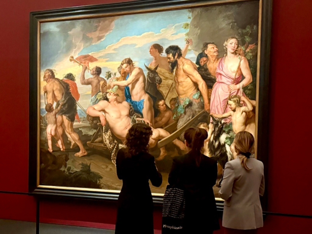 Gemälde mit mehreren teils unbekleideten Figuren in dynamischen Posen vor dramatischem Himmel, drei Personen betrachten das Bild in einem Museum mit roter Wand.