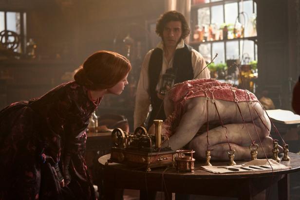 Eine Leiche ohne Kopf kniet auf einem Tisch, ein Teil der Haut ist aufgespannt. Eine Frau begutachtet das, Victor Frankenstein steht im Hintergrund.