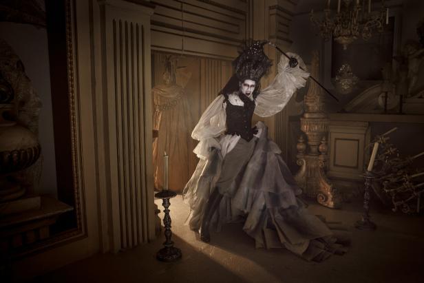 Eine Person in opulenter, historischer Verkleidung mit weiß geschminktem Gesicht und dramatischer Pose steht in einem prunkvollen Raum.